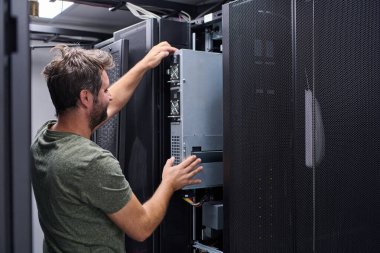 Erkek bilişim teknisyeni veri merkezindeki bir rafa sunucuyu yerleştiriyor, donanım bakımı ve güncellemeleri yapıyor. Modern bir teknolojide teknoloji, takım çalışması, güvenilirlik ve girişim altyapısına odaklanın