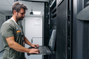 Bir IT teknisyeni, modern bir veri merkezi sunucu odasında sunucuları ve ağ ekipmanlarını yapılandırırken, bakım, izleme ve sistem yönetimi görevlerine odaklanmış bir dizüstü bilgisayarda çalışır..