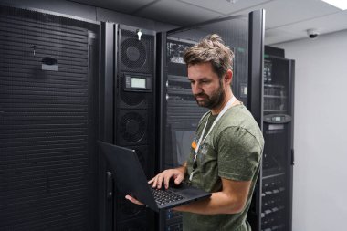 Bilişim teknisyeni, güvenli bir veri merkezinde altyapı sorunlarını çözerken, bir sunucu odasında rafları izlemek, ağ sistemlerini kontrol etmek ve donanım bakımını yapmak için bir dizüstü bilgisayar kullanır..