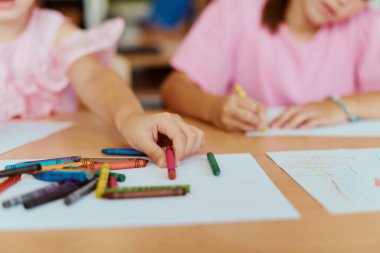 Küçük çocuklar renkli resimler çizmeye odaklanmış bir sınıf masasında boya kalemleri kullanıyorlar. Sahne yaratıcılık, öğrenme, işbirliği ve çocukluk eğlencesini eğitim ortamında aktarıyor.