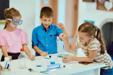 Bir grup çocuk renkli bir sınıf bilim deneyi üzerinde birlikte çalışıyorlar, test tüplerine sıvı döküyorlar ve BTMM becerilerini elle keşif, takım çalışması ve merak yoluyla öğreniyorlar..