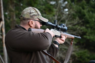 A hunter in a camouflage cap and sunglasses shoulders a scoped rifle in a wooded area, concentrating on a distant target. Outdoor hunting, marksmanship and tactical shooting in nature.