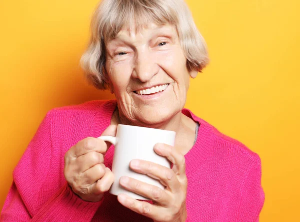 Portrait of old excited lady smiling laughing, holding cup drinking coffee, tea, beverage on yellow background — Stock Photo, Image