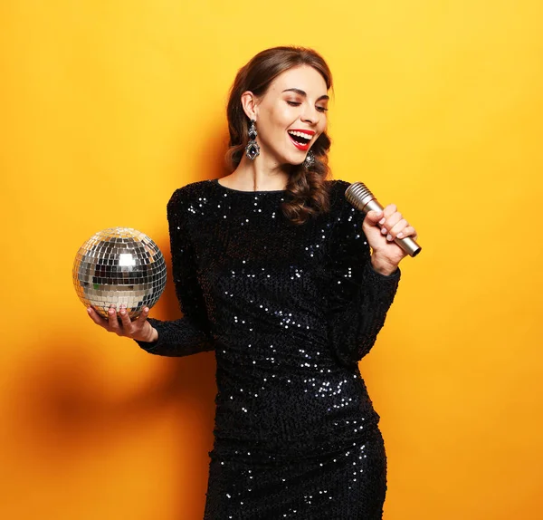 Párty, dovolená a oslavy koncepce: Mladá brunetka s dlouhými kudrnatými vlasy oblečená ve večerních šatech drží mikrofon a disco míč — Stock fotografie