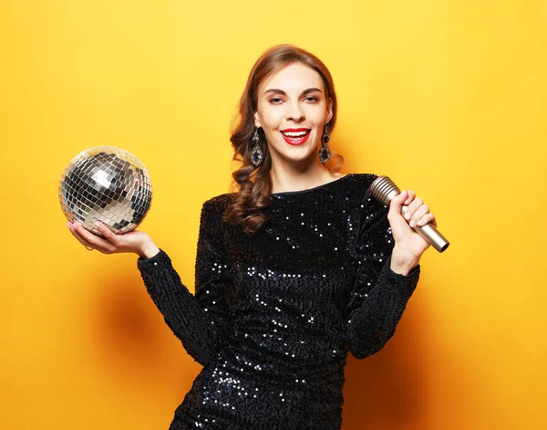 Christmas, celebration party, holiday concept. Young woman in evening dress holding microphone and disco ball. Brighrt make up and Wavy hairstyle.