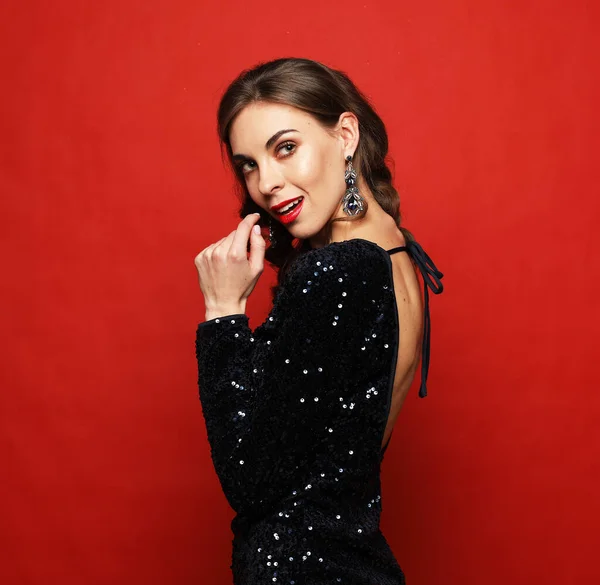 Femme de luxe incroyable en robe de soirée élégante posant sur fond rouge. Yeux fumés lumineux maquillage, lèvres rouges pleines, poils ondulés en bonne santé. — Photo