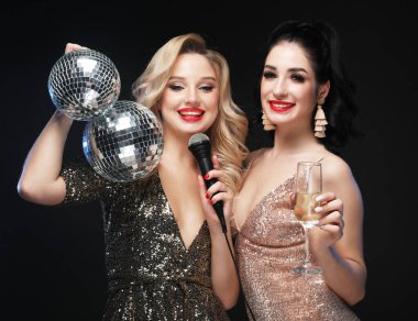 İki güzel kadın, bir sarışın ve parlak elbiseli bir esmer, mikrofonla şarkı söylüyorlar, ellerinde disko topu ve şampanya bardakları var. Parti ve kutlama. Muhteşem saç stilleri ve makyaj..