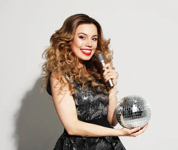 Christmas, celebration party, holiday concept. Young woman in evening dress holding microphone and disco ball. Brighrt make up and Wavy hairstyle.