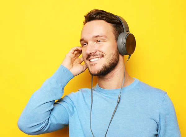 Jóképű férfi kék pulóvert és fejhallgatót visel és mobiltelefont tart sárga alapon. — Stock Fotó