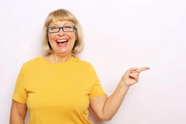 Mosolygó idős nő visel sárga ing és szemüveg felett fehér háttér mutató ujjal — Stock Fotó