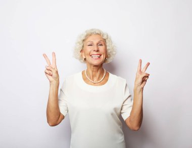 Mutlu, mutlu, gülümseyen, pozitif, yaşlı bir kadın beyaz arka planda V işareti gösteriyor.