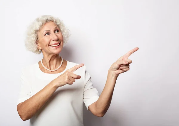 Mujer mayor sonriente con camisa blanca señalando con el dedo el espacio de copia en blanco a un lado — Foto de Stock