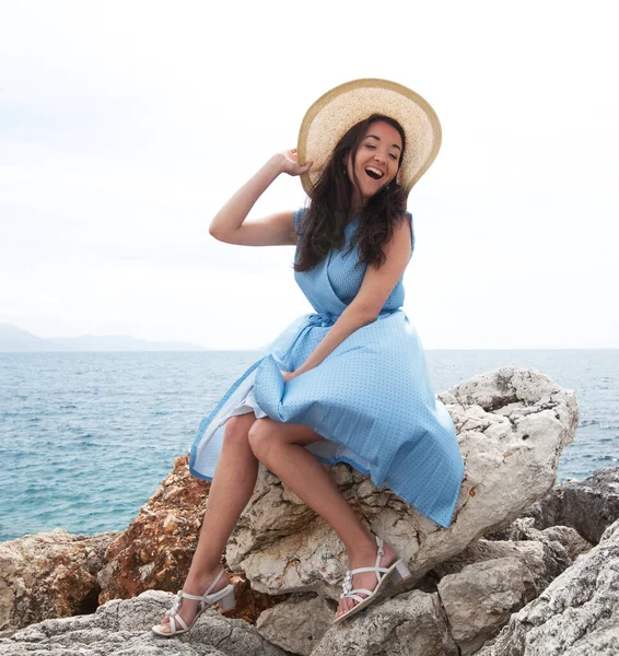 Jovem encantadora com cabelo escuro se senta em uma pedra ao lado do mar. Vestido azul e chapéu de palha. Beleza na moda. — Fotografia de Stock