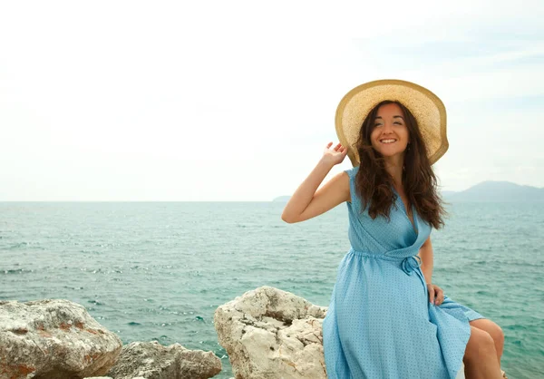 Eine fröhlich lächelnde Brünette in blauem Kleid und Strohhut sitzt auf einem großen Stein, hinter ihr das Meer. Griechenland. Urlaub. — Stockfoto
