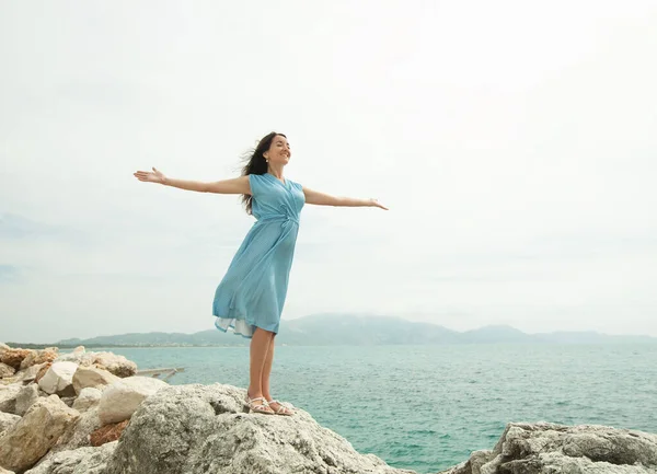 Eine junge Frau mit dunklen Haaren in einem blauen Kleid steht mit ausgestreckten Armen auf einem Stein am Meer — Stockfoto