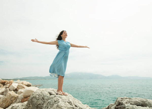 A young woman with dark hair in a blue dress stands on a stone with her arms outstretched, next to the sea, feeling freedom and happiness.