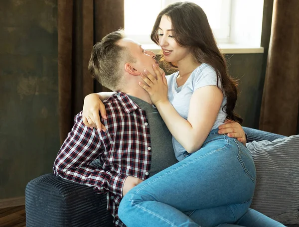 Um par de jovens amantes bonitos estão abraçando dentro de casa no sofá e olhando profundamente nos olhos um do outro com amor — Fotografia de Stock