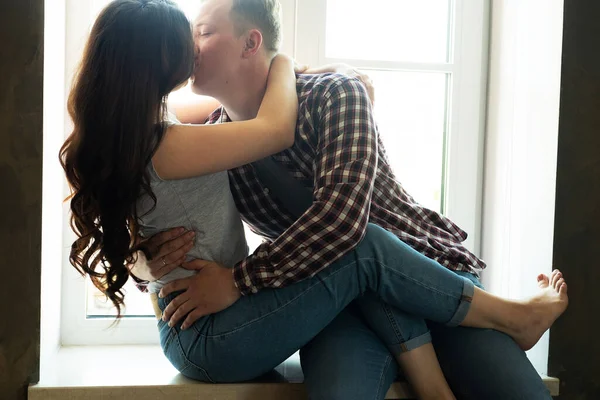 A fiatal pár ölelkezik és csókolózik, miközben az ablakpárkányon ülnek az ablak mellett. — Stock Fotó