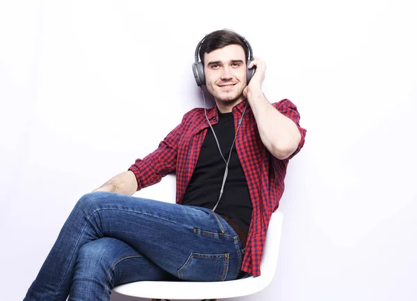 Een knappe jongeman in een geruit hemd en jeans zit in een fauteuil en luistert naar muziek op koptelefoon — Stockfoto