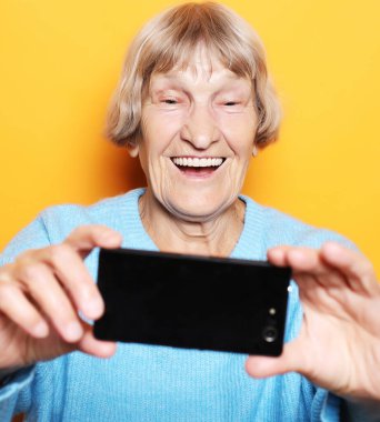 Mavi kazaklı büyükanne gülümsüyor ve sarı arka planda selfie çekiyor.