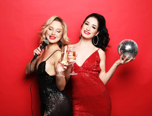 Zwei schöne Frauen, blond und brünett in Kleidern, singen mit einem Mikrofon, halten Discokugeln und ein Glas Wein in der Hand., — Stockfoto