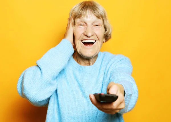 Lifestyle και άνθρωποι έννοια: αστεία γιαγιά κρατά ένα τηλεχειριστήριο τηλεόραση πάνω από κίτρινο φόντο — Φωτογραφία Αρχείου