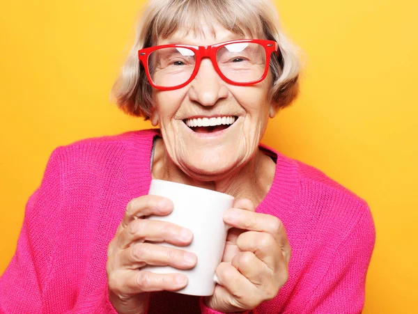 Retrato de velha senhora animada sorrindo rindo, segurando xícara bebendo café, chá, bebida no fundo amarelo — Fotografia de Stock