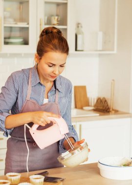 Genç bir kadın pasta için çırpıcı ve krem şanti kullanır, modern mutfakta durur.
