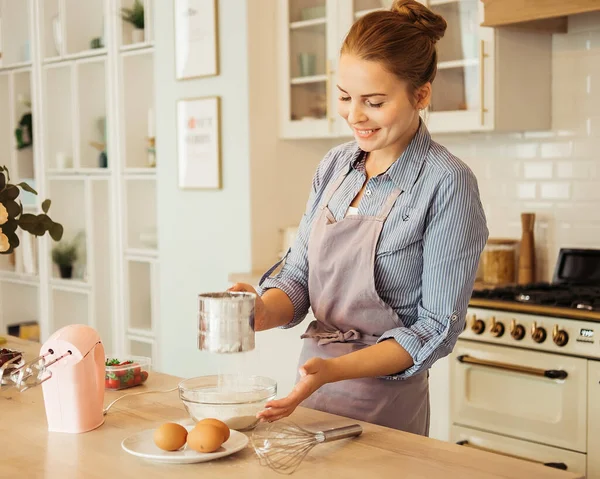 Young woman with toothy smile sifting flour over table before making dough — Stock Photo, Image