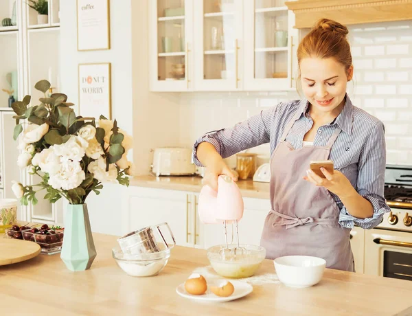 Young woman using mixer standing in kitchen and looking at recipe on mobile phone — Stock Photo, Image