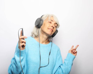 Yaşlı bir kadın beyaz arka planda kulaklıkla müzik dinliyor.