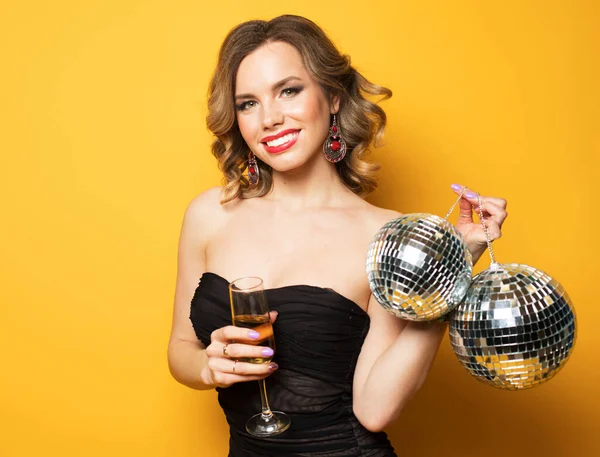 Young blonde woman in evening black dress holding a glass of champagne and disco ball. Brighrt make up and Wavy hairstyle. Over yellow background. — Stock Photo, Image