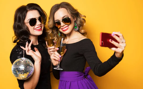 Feier, Party und People-Konzept - zwei modische junge Frauen halten Discokugel in der Hand, trinken Champagner und machen ein Selfie. — Stockfoto