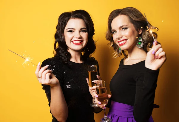 Sarı arka planda şampanya bardakları ve maytapları olan iki genç kadının portresi. — Stok fotoğraf