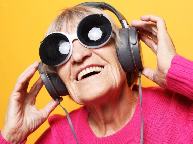Neşeli yaşlı kadın sarı arka planda kulaklıklı müzik dinlerken büyük güneş gözlüğü takıyor. Yaşam tarzı ve insan kavramı.