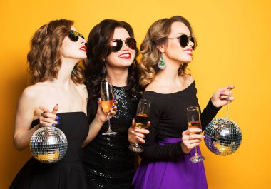 Disko topları olan, şampanya içen ve sarı arka planda eğlenen üç neşeli genç bayan arkadaş. Parti zamanı.
