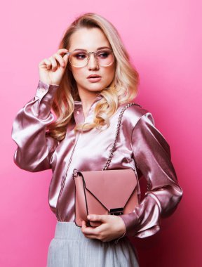 Bluz ve pantolon giymiş güzel sarışın bir kadın. Gözlük takıyor. Elinde pembe arka planda duran bir çanta var..