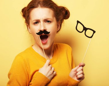 Neşeli, genç, kızıl saçlı kadın elinde parti malzemeleriyle sarı arka planda poz veriyor..