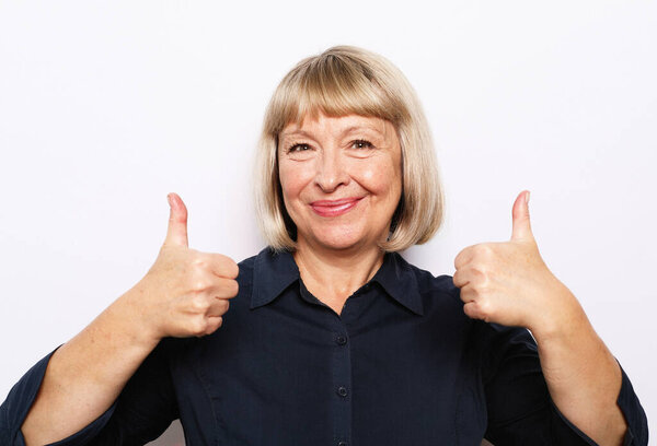Portrait of cheerful old woman in blue shirt showing thumbs up gesture, isolated on white background, close up
