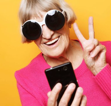 Yaşam tarzı, tehnoloji ve insan konsepti: Yaşlı bir kadın akıllı telefon tutuyor ve V-işareti yapıyor