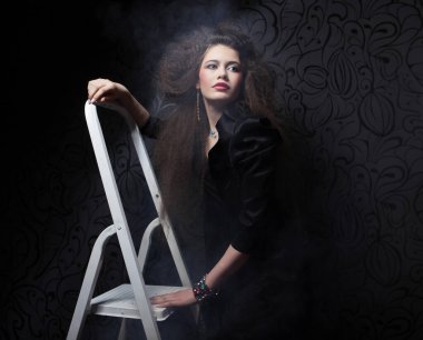 Uzun kıvırcık saçlı, gece makyajlı, siyah elbiseli ve beyaz basamaklı merdivende duran güzel esmer kadın.