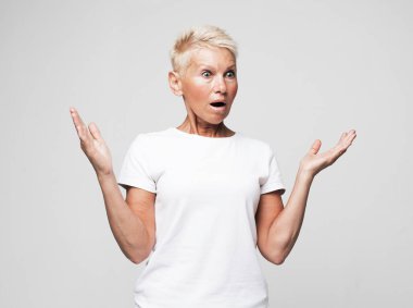 Modern yaşam tarzı, yaşlı insanlar ve duygu konsepti. Yaşlı sarışın, mutlu, mutlu, neşeli, şaşkın, gülümseyen, çığlık atan, yanaklarına el kaldıran, gri arka planda ağzını açan,