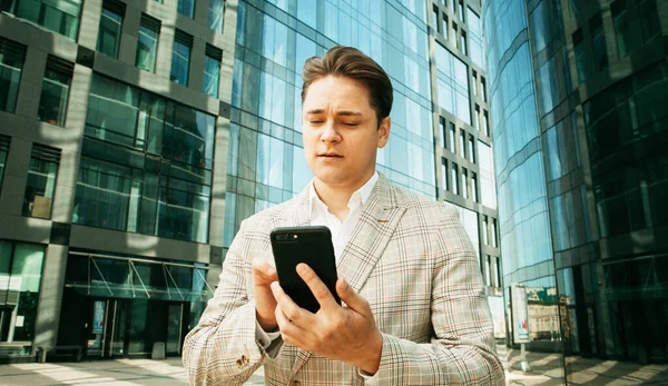 背景に高層ビルが建ち並ぶ都会のオフィスの外で携帯電話のアプリを使っているビジネスマン 仕事のためのスマートフォンを持っている若い白人男性 — ストック写真