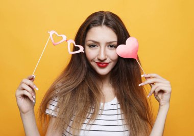 Yaşam tarzı, insanlar ve parti konsepti: Çekici esmer kadın sarı arka planda bir çubukta gözlük ve kırmızı kalp tutuyor.