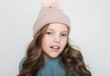 Beyaz arka planda gülüp duran sevimli kız portresi. Mavi kazak ve kış şapkası giyen küçük çocuk gülümseyip kameraya bakıyor..