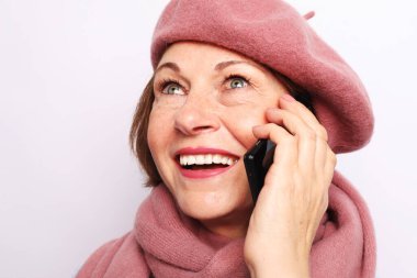 Son sınıf öğrencisi elinde elektronik aletle arkadaşlarıyla telefon görüşmesi yapan yaşlı kadın kendini mutlu hissediyor. Yaşlı kadın büyükannesi beyaz stüdyo geçmişine pembe eşarp ve bere takıyor.