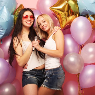 Mikrofonlu iki kadın renkli balonların arka planında karaoke söylüyor.