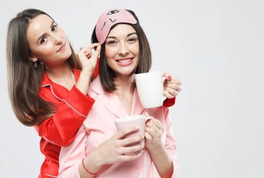 Pijama giymiş, ellerinde fincanlarla sarılıp gülümseyen iki güzel genç kadın..