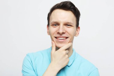 Yakışıklı genç adam gülümsüyor ve gri arka planda çenesine dokunuyor.