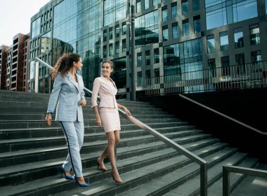 İş dünyası, insanlar ve yaşam tarzı kavramı: İki gülen iş kadını modern ofis binasının merdivenlerinden aşağı inerken konuşuyor.